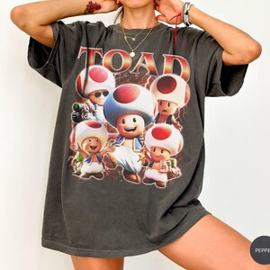 Może przedstawiać: Ciemnoszary t-shirt z krótkim rękawem z dużym nadrukiem graficznym przedstawiającym kilka postaci Toada z serii Super Mario. Słowo TOAD jest nadrukowane pogrubioną, stylizowaną czerwoną i żółtą czcionką nad postaciami, z motywem błyskawic.