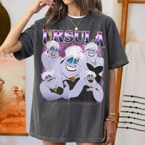 May include: A charcoal gray short-sleeve t-shirt featuring a graphic print of the character Ursula from The Little Mermaid. The design displays multiple illustrations of the purple-skinned sea witch against a purple nebula background with the text URSULA.