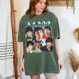 May include: An olive green short-sleeve t-shirt featuring a graphic print of actor Tom Welling. The design includes the text TOM WELLING in a metallic blue font above a collage of four different photographs of the actor in various outfits.