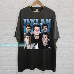 May include: Dark gray t-shirt featuring a graphic of Dylan O'Brien with the name "Dylan O'Brien" in blue text. The shirt has a vintage style and a crew neck.