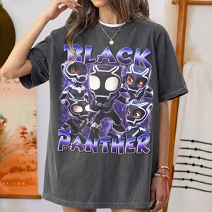 May include: A charcoal gray short-sleeve t-shirt featuring a graphic print of five stylized Black Panther characters in various poses. The text "BLACK PANTHER" is printed in large, bold, purple-outlined letters above and below the characters.