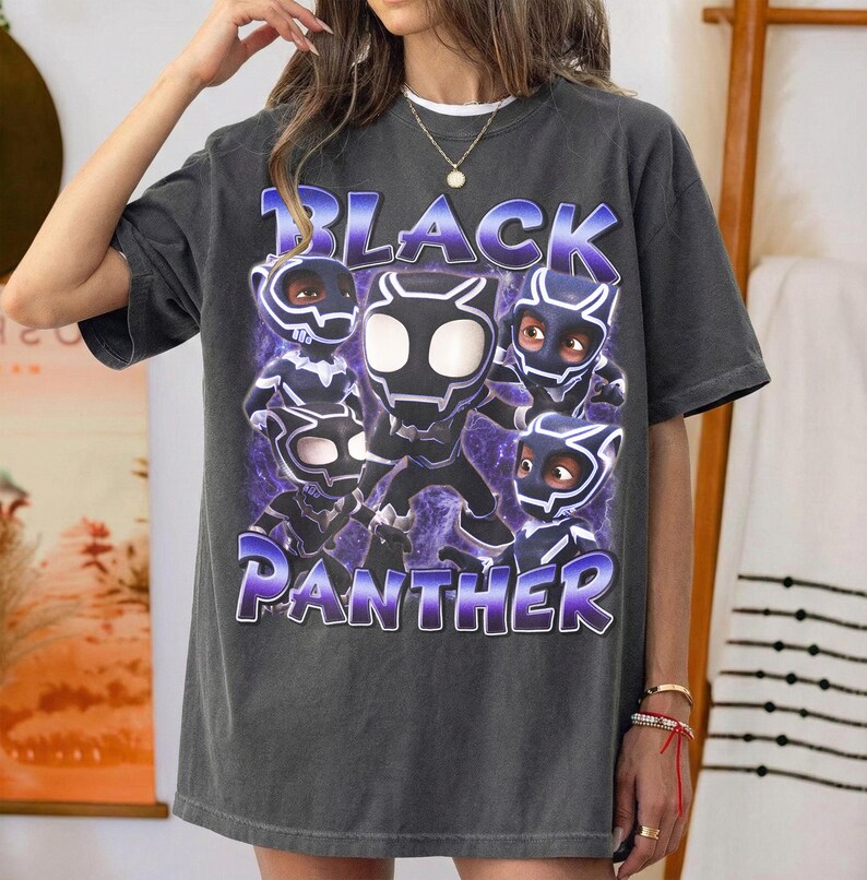 May include: A charcoal gray short-sleeve t-shirt featuring a graphic print of five stylized Black Panther characters in various poses. The text "BLACK PANTHER" is printed in large, bold, purple-outlined letters above and below the characters.