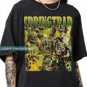 May include: Dark gray t-shirt featuring a graphic of Springtrap characters with a green and yellow color scheme. The word "SPRINGTRAP" is printed in green above the characters. The shirt has a crew neck and short sleeves.