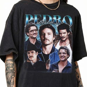 Pode incluir: Uma t-shirt preta de manga curta com uma impressão gráfica do ator Pedro Pascal em vários papéis. O texto "PEDRO Pascal" aparece numa fonte azul metálica no topo. O design inclui cinco retratos fotográficos distintos do ator.