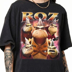 May include: A black short-sleeve t-shirt featuring a graphic print of five identical characters with gray hair, cat-eye glasses, and stern expressions. The text ROZ is printed in large gold letters above the characters, with CDA 001 on a vest.