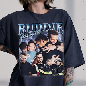 May include: A charcoal grey short-sleeve t-shirt featuring a graphic print with the text "BUDDIE" and "Buck and Eddie" above a collage of images showing two men embracing and wearing firefighter uniforms with reflective yellow and silver stripes.