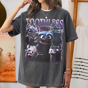 May include: A charcoal gray short-sleeve t-shirt featuring a graphic print of five black dragon characters from the movie Toothless. The word TOOTHLESS is printed in large purple and white letters above the dragons, set against a purple lightning background.