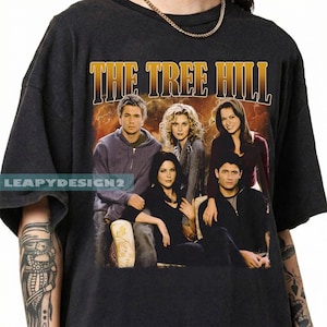 May include: Dark gray t-shirt featuring a group photo of actors from the television show "The Tree Hill" with the show's title in gold lettering. The shirt has a vintage look with a faded graphic.