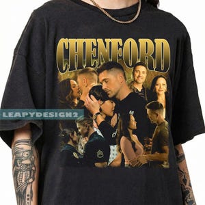May include: Black t-shirt featuring a group of people with the word "CHENFORD" in gold lettering. The shirt has a vintage style design with a collage of images of people. The brand name "LEAPYDESIGN2" is printed on the left side.