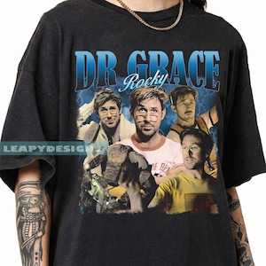 Puede incluir: Camiseta negra con un diseño gráfico con el texto "DR GRACE Rocky" en azul y blanco. El diseño incluye múltiples imágenes de una persona con gafas. La camiseta tiene mangas cortas.