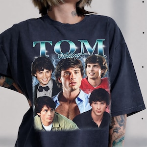 May include: A charcoal gray short-sleeve t-shirt featuring a graphic print of actor Tom Welling. The design includes the text "TOM Welling" in a metallic blue font above a collage of five different photographs of the actor in various outfits.