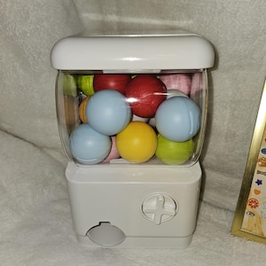 May include: A white gumball machine with a clear, rounded container filled with colorful gumballs. The gumballs are various colors, including red, blue, yellow, and green. The machine has a white top and a dispensing slot at the bottom.
