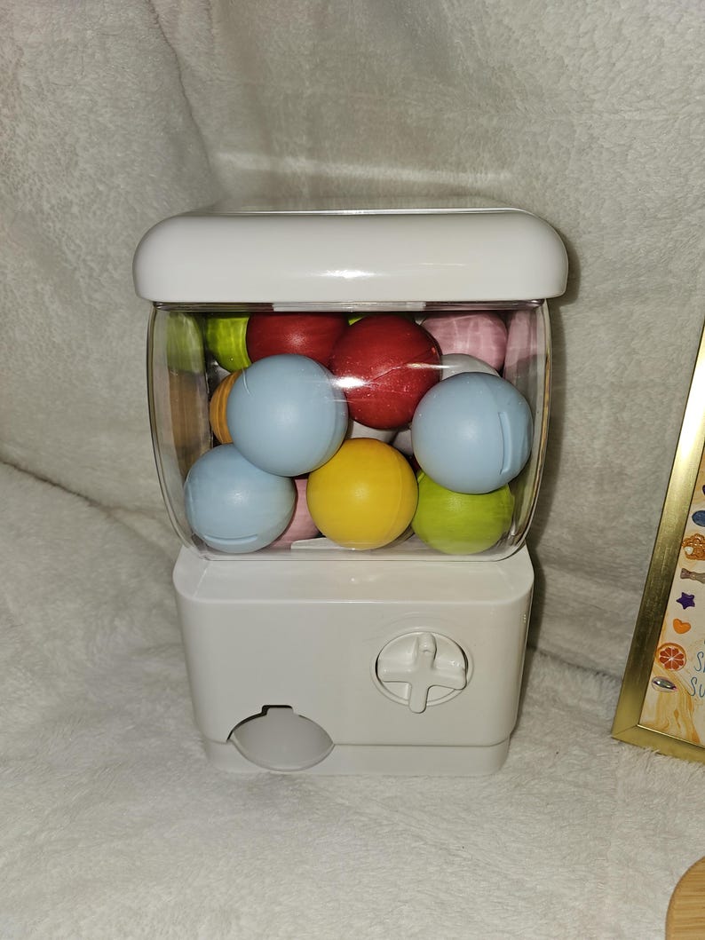 May include: A white gumball machine with a clear, rounded container filled with colorful gumballs. The gumballs are various colors, including red, blue, yellow, and green. The machine has a white top and a dispensing slot at the bottom.