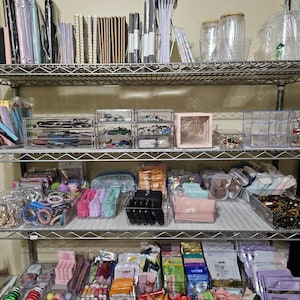 May include: A multi-tiered metal shelf filled with various items. The top shelf holds notebooks, pens, and jars. The middle shelf contains clear organizers with jewelry and accessories. The bottom shelf displays beauty products and colorful items.