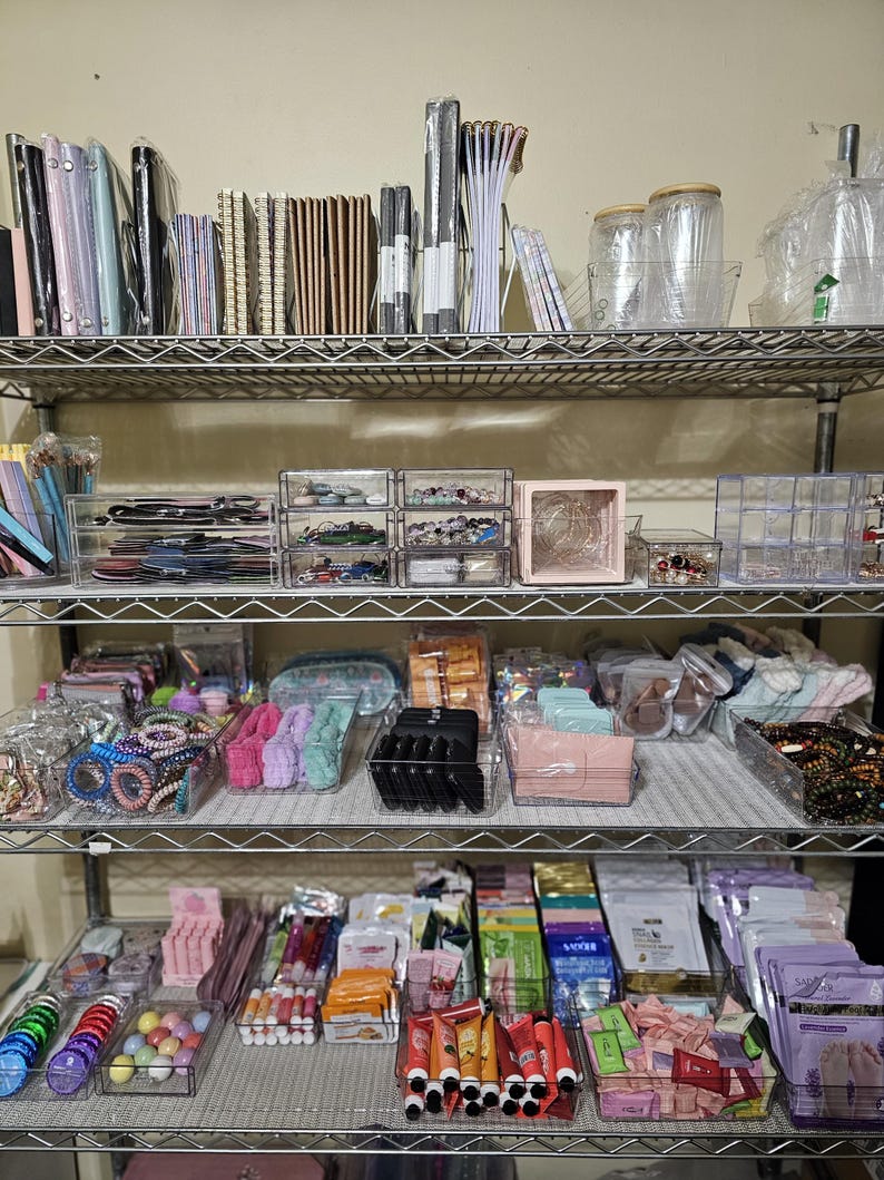 May include: A multi-tiered metal shelf filled with various items. The top shelf holds notebooks, pens, and jars. The middle shelf contains clear organizers with jewelry and accessories. The bottom shelf displays beauty products and colorful items.