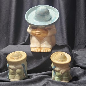 May include: A ceramic figurine of a man wearing a sombrero, with two matching salt and pepper shakers. The main figure is in a seated position, with a light blue sombrero and a white outfit. The salt and pepper shakers are smaller versions of the main figure.
