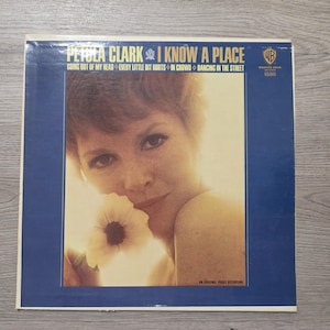 Peut inclure: Pochette de disque vinyle vintage de Petula Clark, intitulée "I Know a Place". La couverture, avec un cadre bleu, présente une photo de Petula Clark tenant une fleur. Les titres des chansons sont listés. Le logo Warner Bros. est visible.