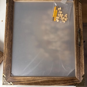 May include: A rectangular wooden frame with a clear plastic sheet inside. The frame has decorative corner accents and a handle. A small plastic bag contains hardware, including yellow anchors and small wooden pegs.