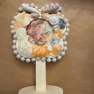 May include: A handcrafted photo frame decorated with seashells and faux pearls. The frame surrounds a circular photo of a mother and child. The frame is mounted on a wooden stand, perfect for displaying on a desk or shelf.