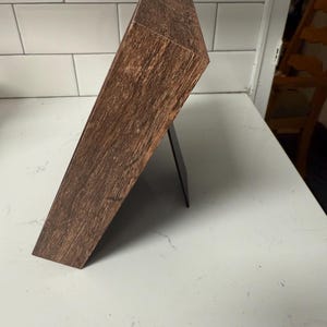 以下が含まれることがあります： 黒いスタンドに立てかけられた、濃い茶色の木製フォトフレーム。フレームは木目調の質感で、白い表面に置かれています。フレームは写真やアートワークを収めるように設計されています。