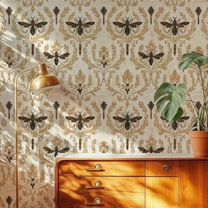 Op de afbeelding: Een houten dressoir met drie lades en een kastdeur, geplaatst tegen behang met een herhalend bijen- en damastpatroon. Links staat een goudkleurige vloerlamp en bovenop staat een monstera-plant in een pot.