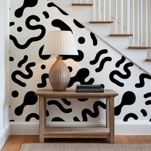 May include: A wooden console table with a lower shelf, topped with a ribbed ceramic table lamp featuring a white pleated shade. The wall behind the table is covered in white wallpaper with a repeating pattern of black organic shapes and dots.