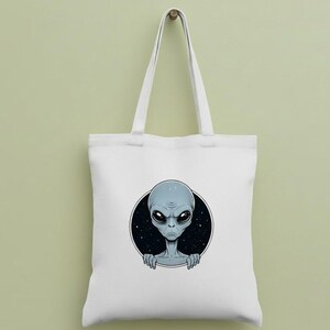 May include: White tote bag featuring a gray alien graphic within a black circle against a starry background. The alien has large black eyes and is peeking out from behind the circle. The bag has two white handles.