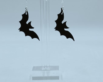 May include: A pair of black bat-shaped earrings. The earrings are hanging from silver-colored hooks. The bats have a detailed wing design. The earrings are displayed on a clear acrylic stand against a white background.
