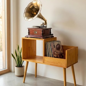 May include: A vintage gramophone with a brass horn sits atop a wooden cabinet with record storage. The cabinet is made of light-colored wood and has a mid-century modern design. A potted plant is visible in the background.