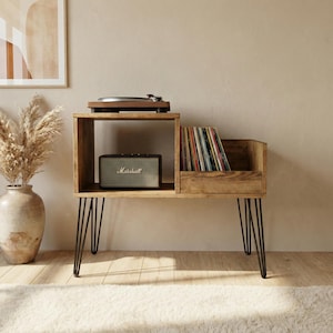 Puede incluir: Soporte de tocadiscos de madera con un tocadiscos en la parte superior, un altavoz Marshall y una sección para discos de vinilo. El soporte tiene patas de horquilla negras y un acabado de madera natural. El soporte mide 76 cm de ancho.