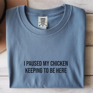 Könnte beinhalten: Hellblaues T-Shirt mit dem schwarzen Text "I PAUSED MY CHICKEN KEEPING TO BE HERE". Das Shirt hat einen Rundhalsausschnitt und ein Etikett "COMFORT COLORS".