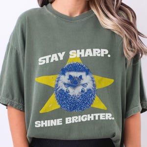 Puede incluir: Camiseta verde oliva con un erizo azul dentro de una estrella amarilla. El texto "STAY SHARP." está encima del erizo, y "SHINE BRIGHTER." debajo. La camiseta tiene un aspecto vintage y desgastado.