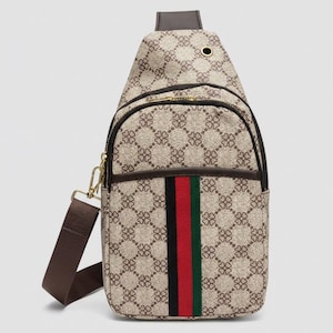 May include: A tan sling bag with a repeating floral pattern. The bag has a brown adjustable strap and a vertical stripe of red, black, and green. The bag has a zippered main compartment and a smaller front pocket.