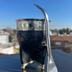 Puede incluir: Un recipiente de yerba mate negro con borde plateado y detalles decorativos. Una pajita bombilla plateada descansa junto al recipiente. El recipiente tiene una forma única con tres pequeños pies dorados.