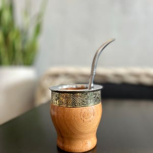 May include: A wooden yerba mate gourd with a silver rim and a decorative band featuring an embossed floral design. A stainless steel bombilla straw is inserted into the gourd. The gourd sits on a dark surface.