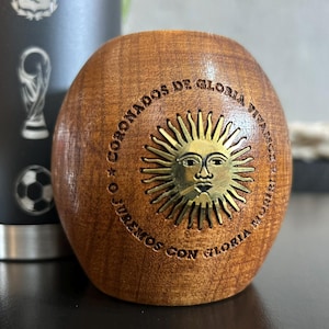 Pu&ograve; includere: Una tazza di mate in legno con un emblema solare in ottone e testo inciso. La tazza &egrave; di forma ovale e presenta venature naturali del legno. L'emblema in ottone raffigura un sole con un volto. Il testo recita "CORONADOS DE GLORIA VIVAMOS O JUREMOS CON GLORIA MORIR".