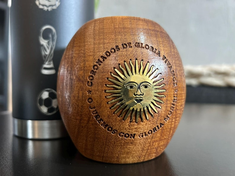 Pu&ograve; includere: Una tazza di mate in legno con un emblema solare in ottone e testo inciso. La tazza &egrave; di forma ovale e presenta venature naturali del legno. L'emblema in ottone raffigura un sole con un volto. Il testo recita "CORONADOS DE GLORIA VIVAMOS O JUREMOS CON GLORIA MORIR".