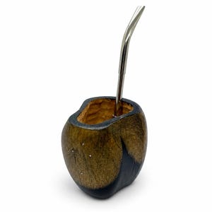 Może przedstawiać: Ciemnobrązowe i czarne drewniane naczynie do yerba mate o naturalnej, teksturowanej powierzchni oraz gładkiej, zakrzywionej krawędzi. Smukła, polerowana metalowa rurka bombilla w kolorze srebrnym jest umieszczona w otworze naczynia.