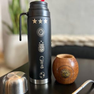 Pu&ograve; includere: Borraccia isolante nera con manico, con simboli incisi tra cui stelle, un sole, un pallone da calcio e un trofeo. Include un coperchio in acciaio inossidabile, una tazza di legno e una cannuccia di metallo.