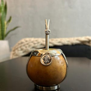 Puede incluir: Un mate de calabaza redondo de color tostado con un acabado liso y natural, encajado en un marco de metal plateado pulido. Una bombilla de metal plateado a juego, con un pequeño emblema de flor de lis, descansa dentro del recipiente.