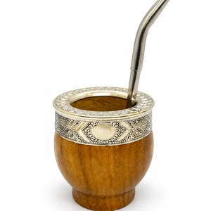 Può includere: Un contenitore per mate in legno con base arrotondata e finitura liscia color marrone chiaro. Il bordo superiore presenta un'ampia fascia metallica decorativa color argento con incisioni geometriche e circolari. Una cannuccia in metallo.