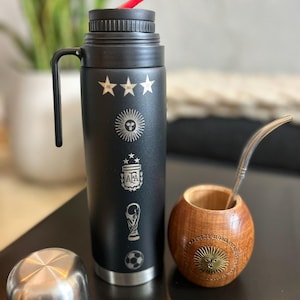 Pu&ograve; includere: Un thermos nero con simboli calcistici argentini incisi e una cannuccia rossa. Accanto, una zucca di legno con cannuccia in metallo e coperchio argentato. La zucca ha un emblema solare decorativo.