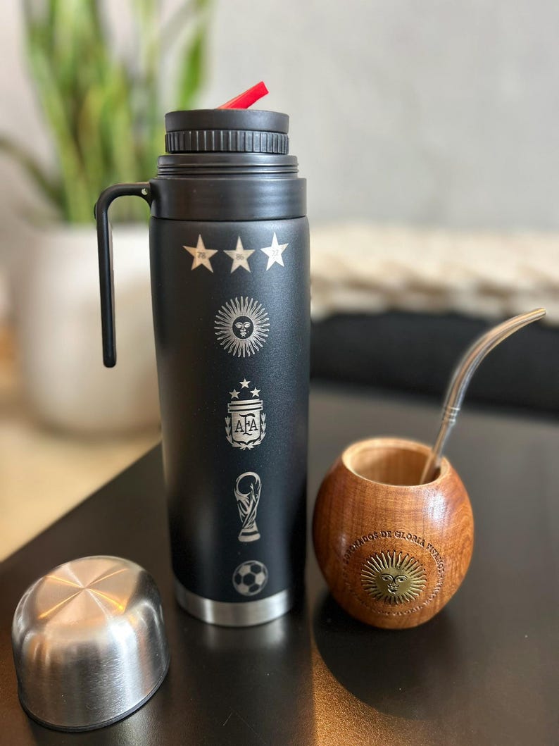 Pu&ograve; includere: Un thermos nero con simboli calcistici argentini incisi e una cannuccia rossa. Accanto, una zucca di legno con cannuccia in metallo e coperchio argentato. La zucca ha un emblema solare decorativo.