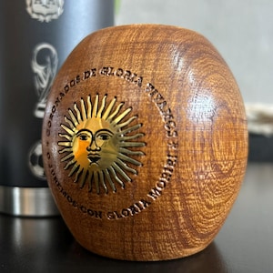 Pu&ograve; includere: Una tazza di mate in legno con un disegno a forma di sole e la scritta "CORONADOS DE GLORIA VIVAMOS CON GLORIA MORIR". La tazza &egrave; marrone con un emblema solare dorato. Sullo sfondo &egrave; visibile un bicchiere nero.