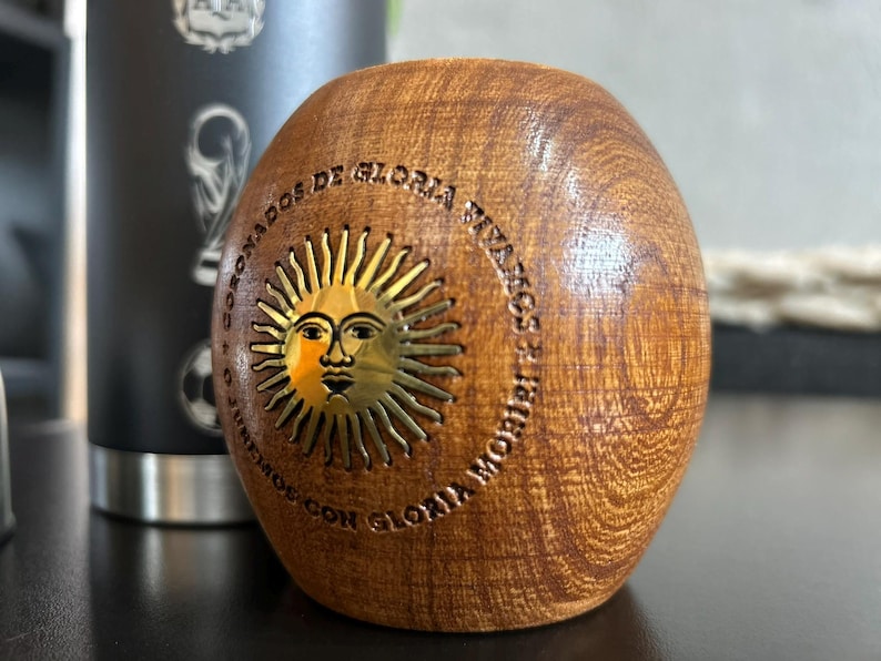 Pu&ograve; includere: Una tazza di mate in legno con un disegno a forma di sole e la scritta "CORONADOS DE GLORIA VIVAMOS CON GLORIA MORIR". La tazza &egrave; marrone con un emblema solare dorato. Sullo sfondo &egrave; visibile un bicchiere nero.