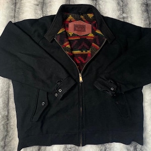 Puede incluir: Chaqueta negra con cierre de cremallera completo y puños abotonados. El forro interior presenta un colorido estampado geométrico en rojo, amarillo y verde. Un parche de cuero marrón con texto es visible en el cuello interior.