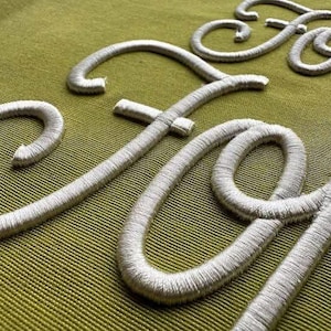 May include: Close-up of silver embroidered lettering on a green fabric background. The letters appear to spell out a word or phrase, with a focus on the detailed stitching and texture of the embroidery.