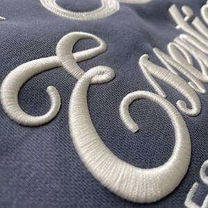 May include: Close-up of a blue fabric with white embroidered text. The embroidery is thick and textured, with a cursive font. The letters are large and stand out against the blue background. The overall effect is elegant and detailed.