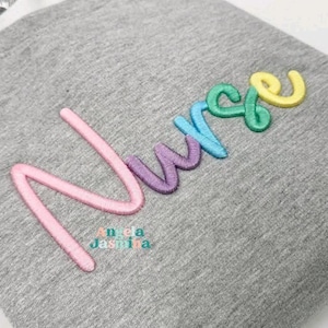 May include: A gray sweatshirt with the embroidered word "Nurse" in pastel colors: pink, purple, blue, green, and yellow. The text "Angela Jashina" is embroidered below the word "Nurse".