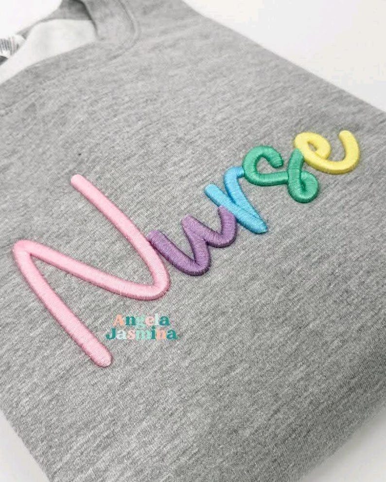 May include: A gray sweatshirt with the embroidered word "Nurse" in pastel colors: pink, purple, blue, green, and yellow. The text "Angela Jashina" is embroidered below the word "Nurse".